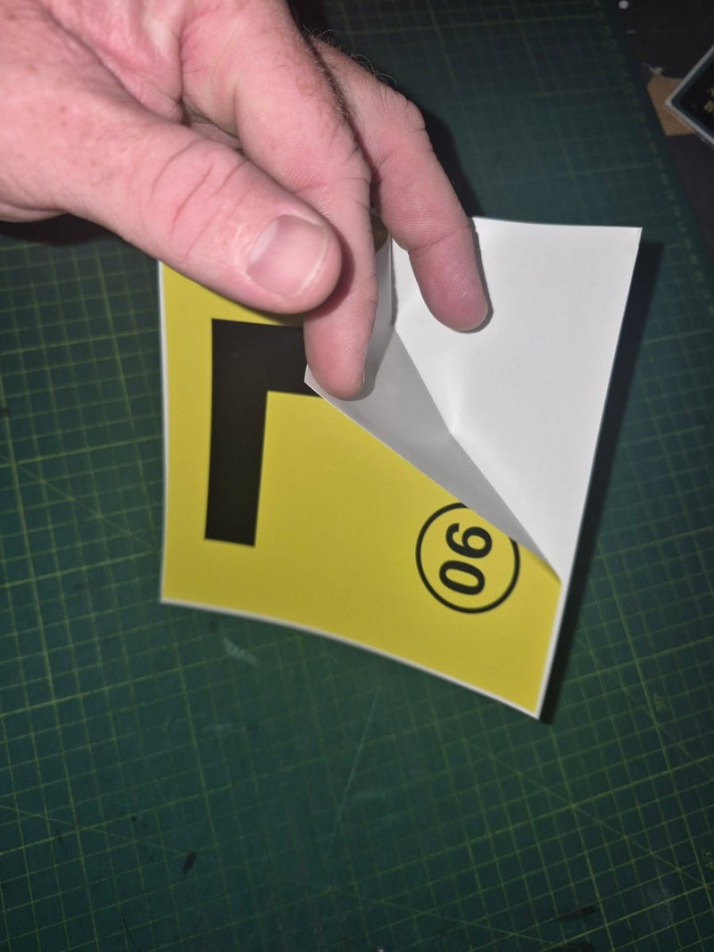 Hand holding a yellow sign with a black letter 'L' and number '90' on a green cutting mat.