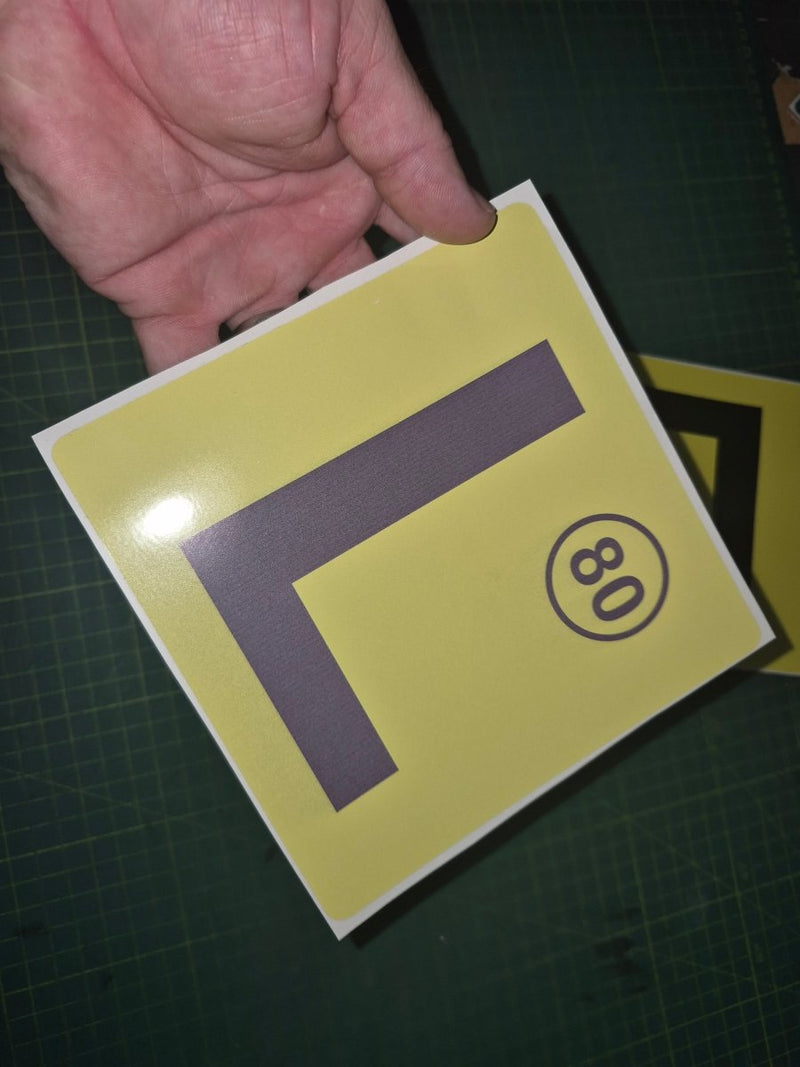 Yellow decal with a large 'L' and number 80 held by a hand on a dark surface.