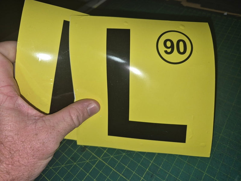 Yellow sign with a black 'L' and number 90 held by a hand on a green cutting mat.