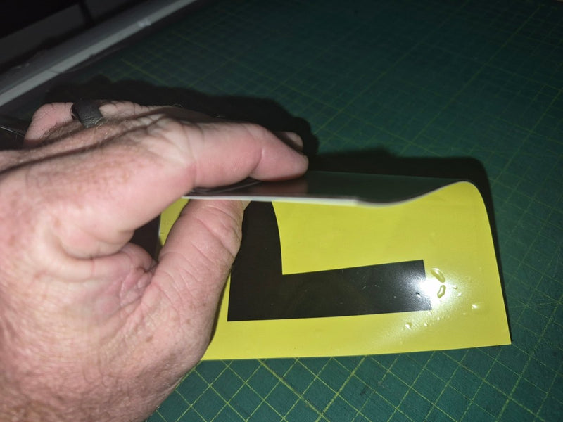 Hand holding a black 'L' plate on a yellow background with a green cutting mat.