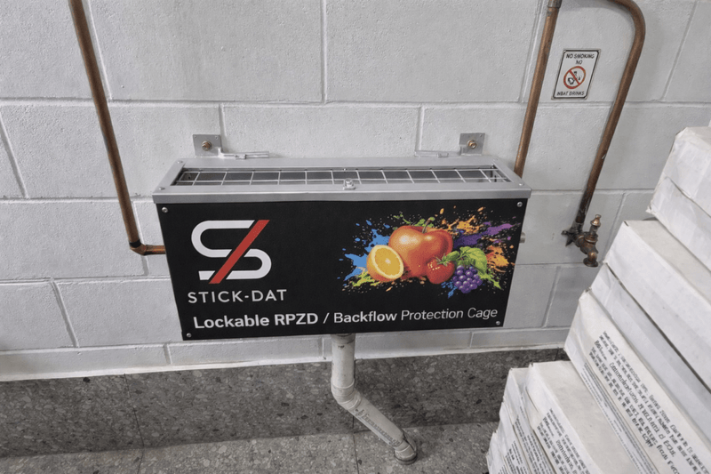 Lockable RPZD/backflow protection cage with Stick-Dat branding on a tiled wall.