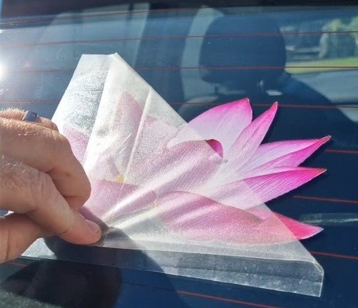 Hand holding a pink flower-shaped car decal against a car window.