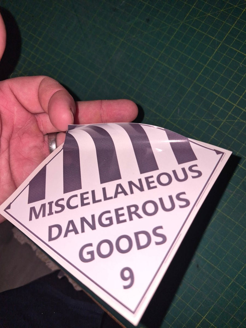 Hand holding a triangular label with 'Miscellaneous Dangerous Goods' text on a green cutting mat.