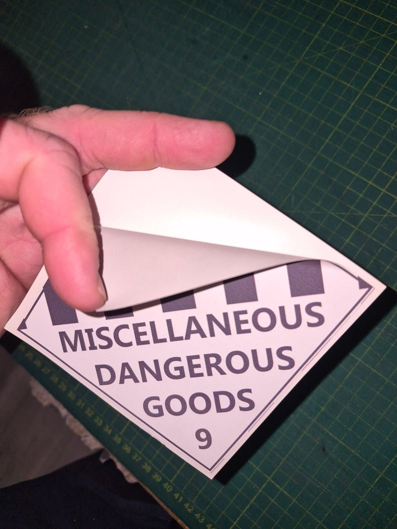 Hand holding a triangular label with 'Miscellaneous Dangerous Goods' text on a green cutting mat.