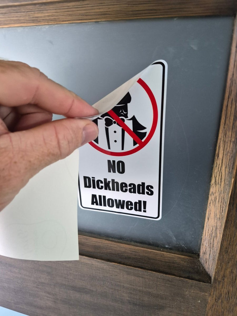 Hand holding a sticker with 'No Dickheads Allowed!' text against a mirror.