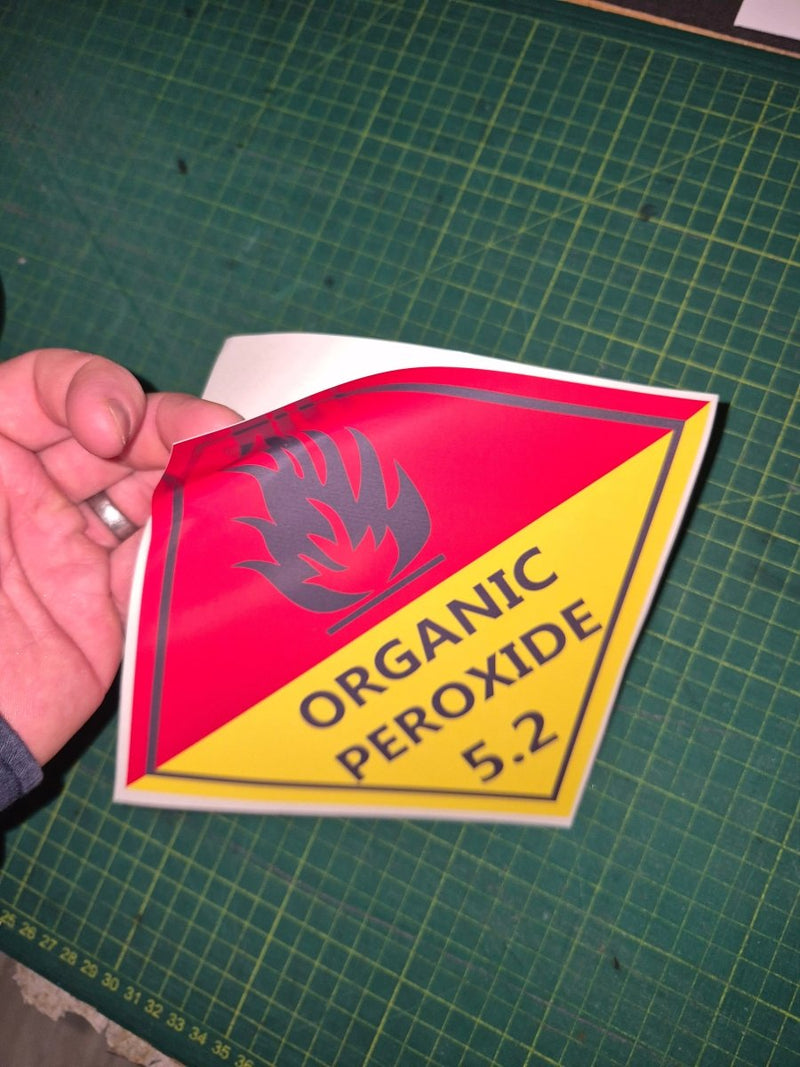 Organic Peroxide 5.2 Sticker Hand holding a red and yellow warning label on a green cutting mat