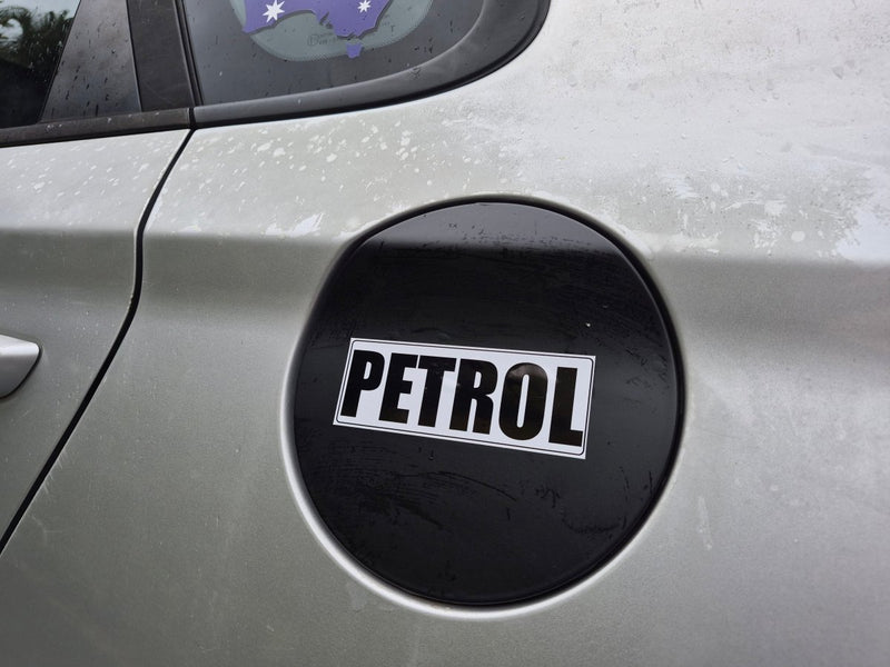A close-up of a black oval fuel door sticker with the word 'PETROL' in bold white letters on a silver car body.