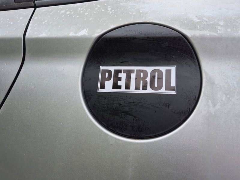 A close-up of a silver car fuel door with a black circular emblem featuring the word 'PETROL' in white letters.