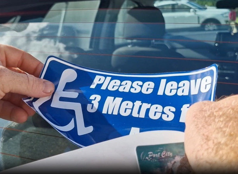 Hand holding a blue and white decal with a wheelchair icon and text 'Please leave 3 Metres' in front of a car.