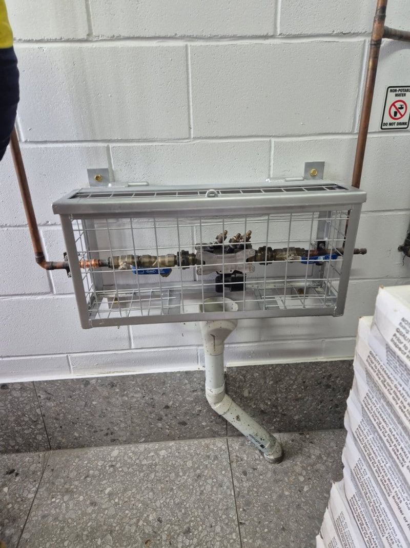 Plumbers cage attached to a wall with pipes and a stack of newspapers on the ground.