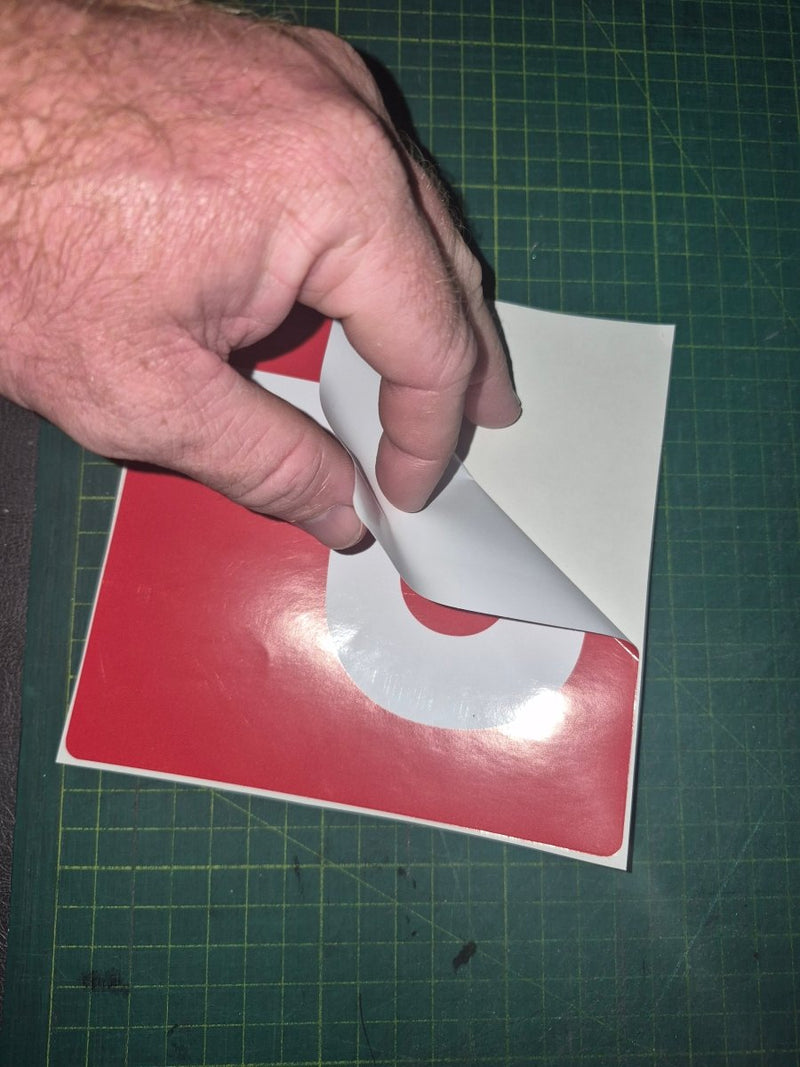 Hand peeling a white sticker from a red and white square on a green cutting mat.