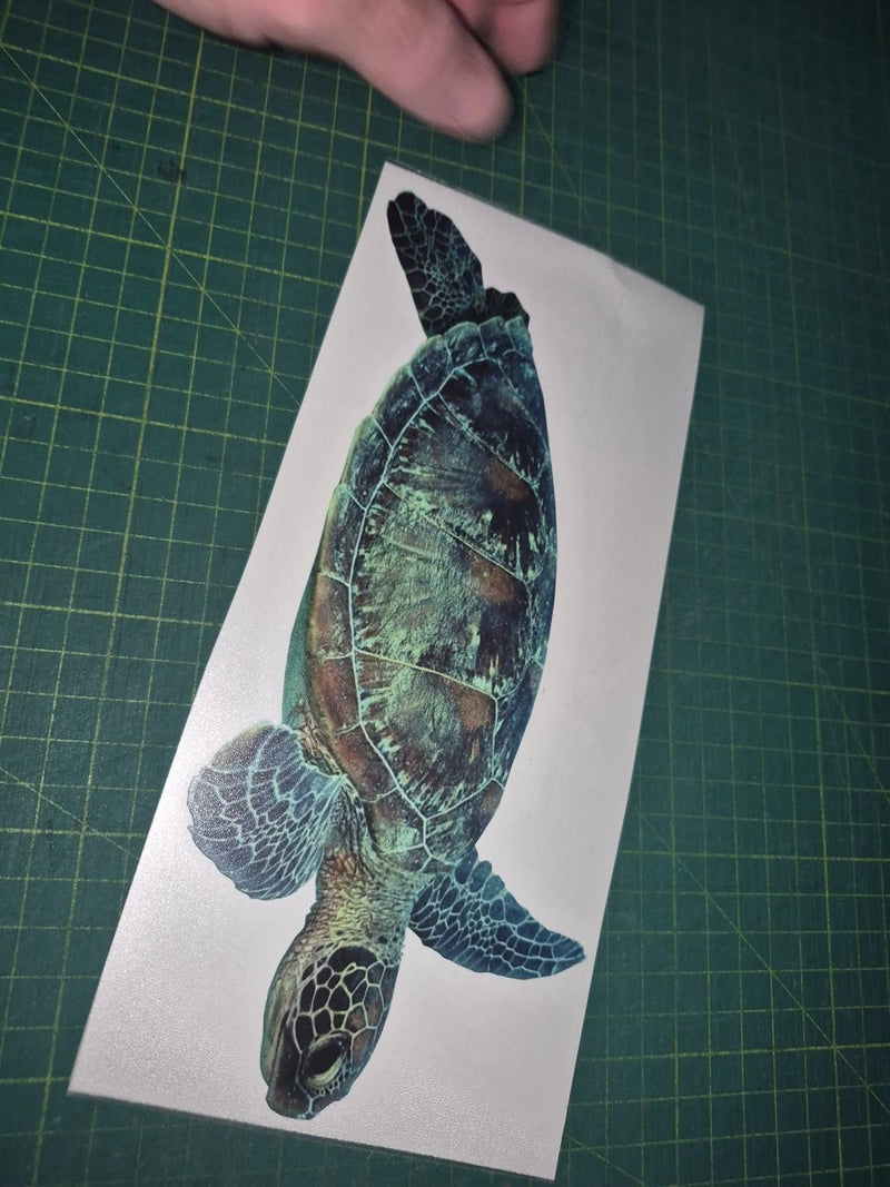 Sticker of a turtle on a piece of paper held by a hand on a green cutting mat.