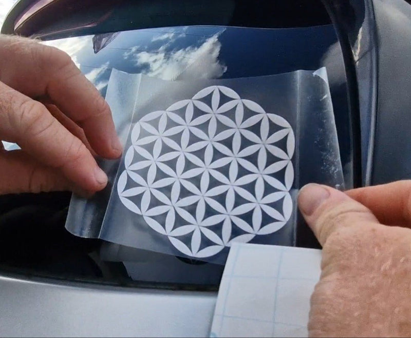 Person applying a white flower of life decal to a car window, geometric design sticker