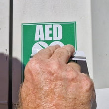 Load image into Gallery viewer, Hand pressing a button on an AED machine with a green sign above it.
