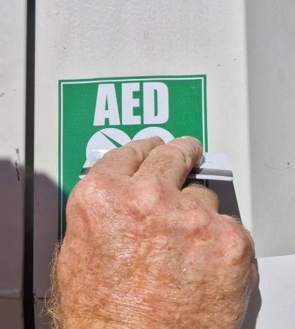 Hand pressing a button on an AED machine with a green sign above it.