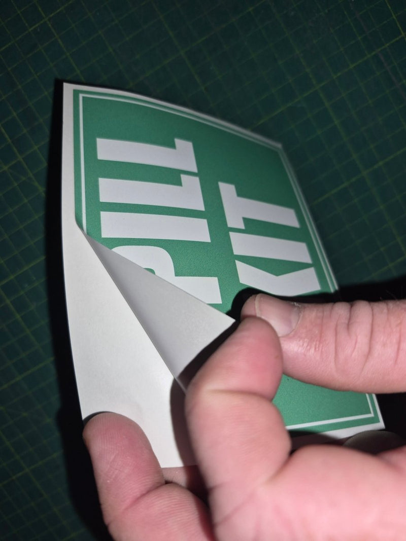 Hand holding a green and white 'Spill Kit' sign on a grid background