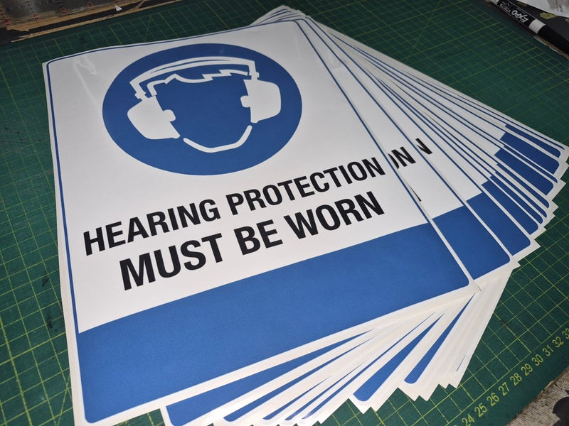 Stack of 'Hearing Protection Must Be Worn' signs on a green cutting mat.