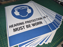 Load image into Gallery viewer, Stack of 'Hearing Protection Must Be Worn' signs on a green cutting mat.
