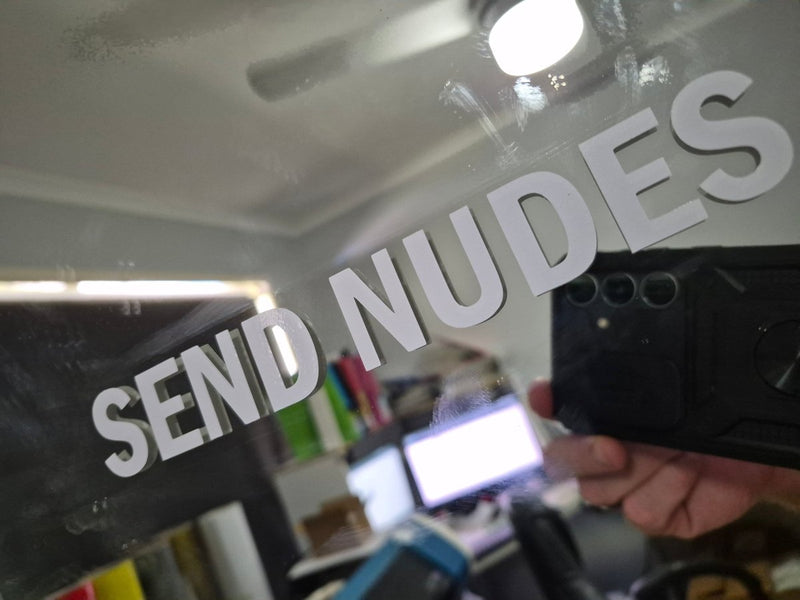 Sticker Text 'SEND NUDES' overlaid on a blurred indoor setting with electronic devices.