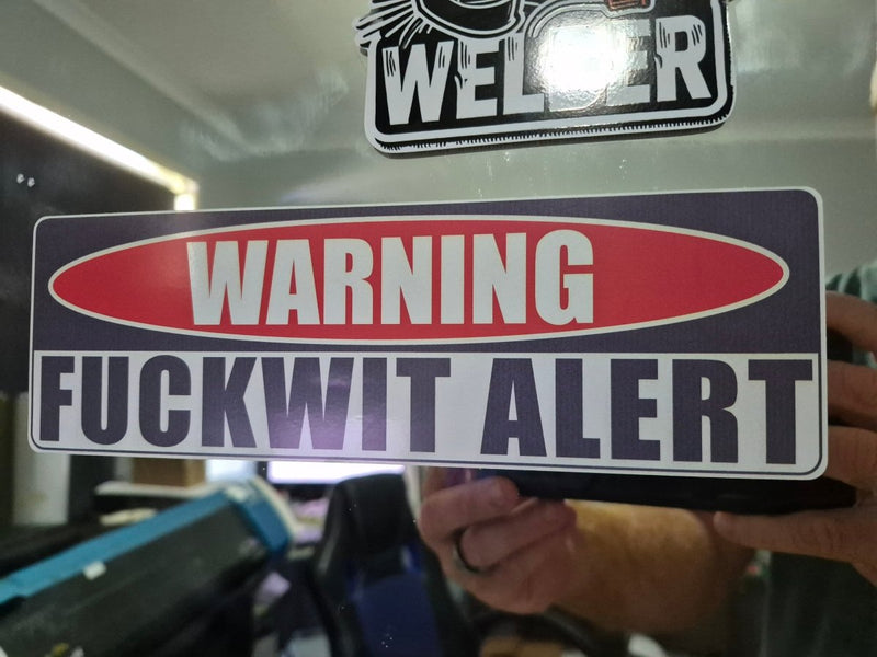 Sticker with 'WARNING FUCKWIT ALERT' text held by a person, with a blurred background.