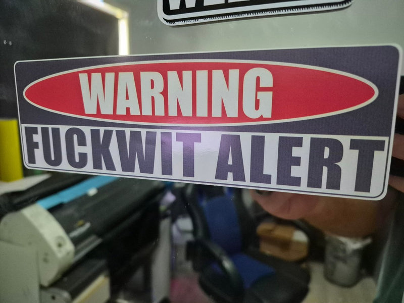 Warning sticker with text 'WARNING FUCKWIT ALERT' held in front of a machine.