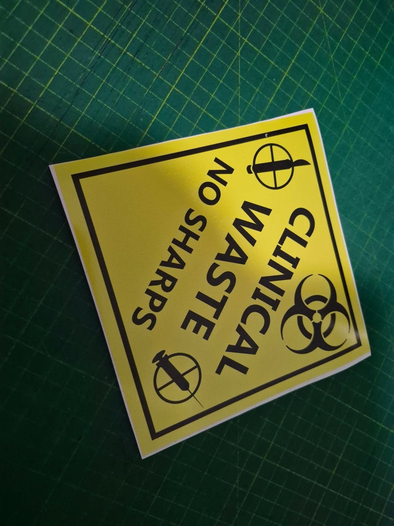 Yellow warning label with 'Clinical Waste No Sharps' text on a green cutting mat.