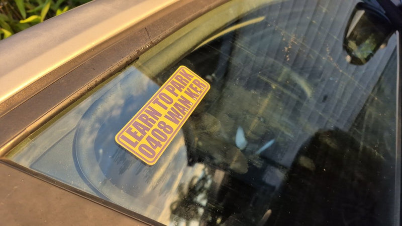 Yellow sticker on a car window with text 'LEARN TO PARK WANKER'.