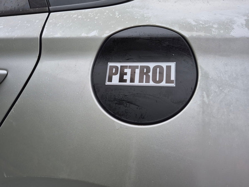A close-up of a silver car fuel door with a black circular cover featuring a white sticker that reads 'PETROL'.